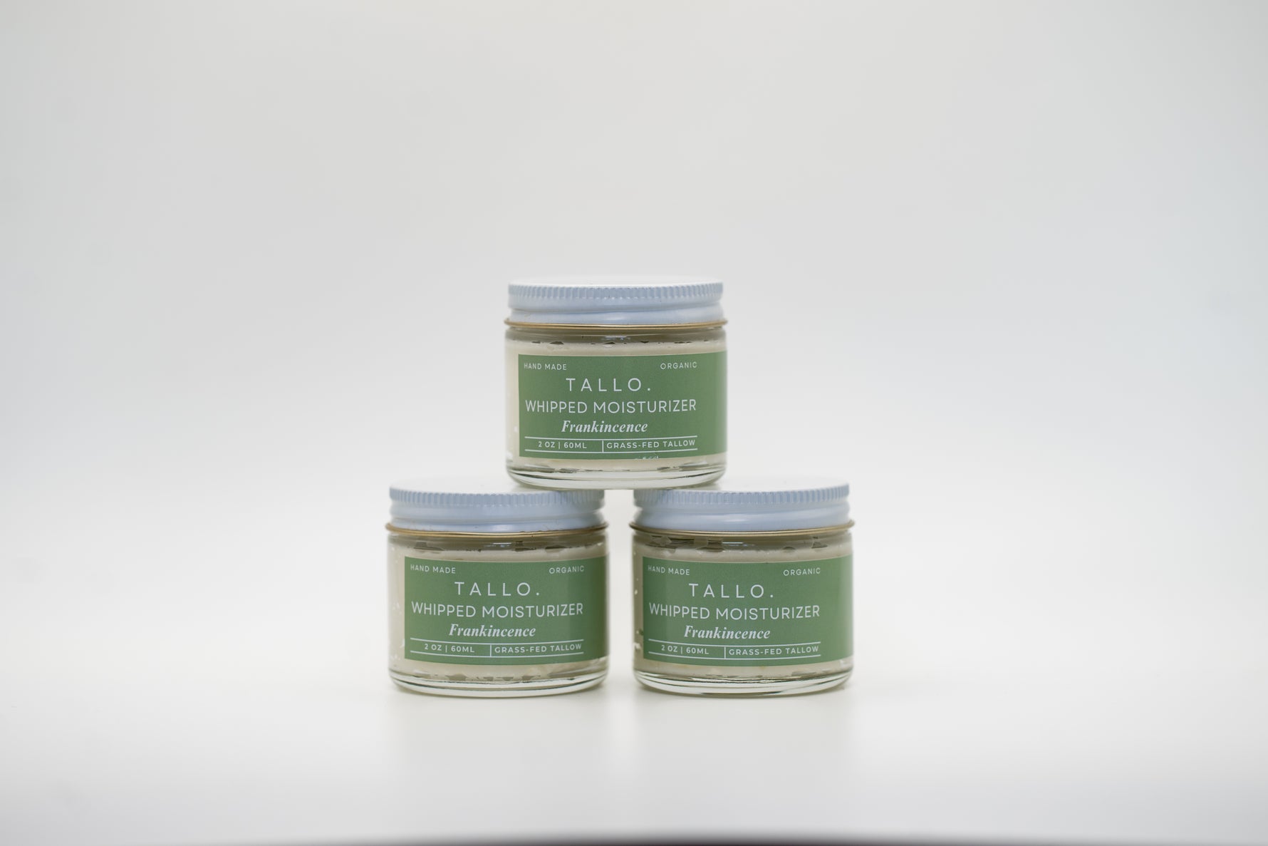 A set of three small glass jars of Tallo Whipped Moisturizer with green labels and white lids, stacked in a pyramid formation. The labels read Frankincense. The product is labeled as handmade, organic, and made with grass-fed tallow.
