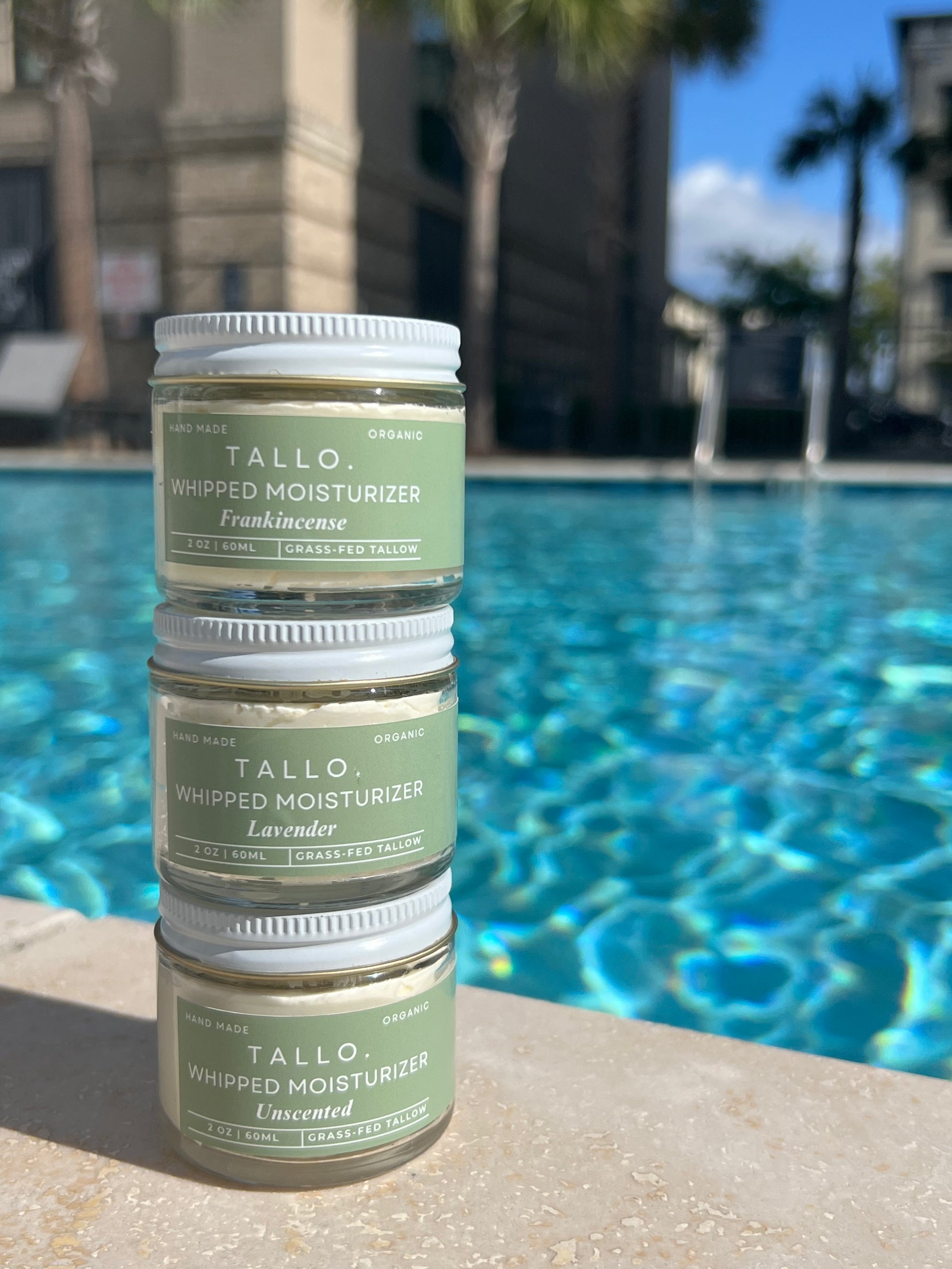 A set of three small glass jars of Tallo Whipped Moisturizer with green labels and white lids, stacked on top of each other. The labels indicate different variations: Frankincense, Lavender, and Unscented. The product is labeled as handmade, organic, and made with grass-fed tallow.