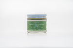 A small glass jar of skincare product with a green label and blue lid. The label lists natural ingredients including grass-fed beef tallow, cold-pressed castor oil, local honey, jojoba oil, and essential oils. It also includes application instructions to apply to face and body as needed, and displays the website GETTALLO.COM and Instagram handle @GETTALLO.