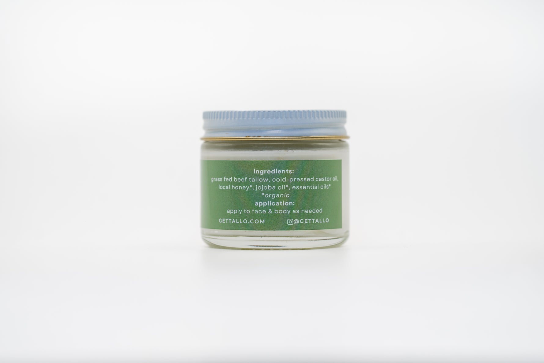 A small glass jar of skincare product with a green label and blue lid. The label lists natural ingredients including grass-fed beef tallow, cold-pressed castor oil, local honey, jojoba oil, and essential oils. It also includes application instructions to apply to face and body as needed, and displays the website GETTALLO.COM and Instagram handle @GETTALLO.