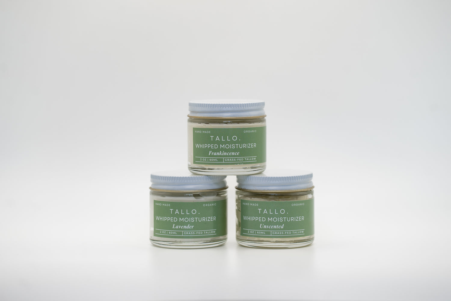 A set of three small glass jars of Tallo Whipped Moisturizer with green labels and white lids, stacked in a pyramid formation. The labels indicate different variations: Frankincense, Lavender, and Unscented. The product is labeled as handmade, organic, and made with grass-fed tallow.