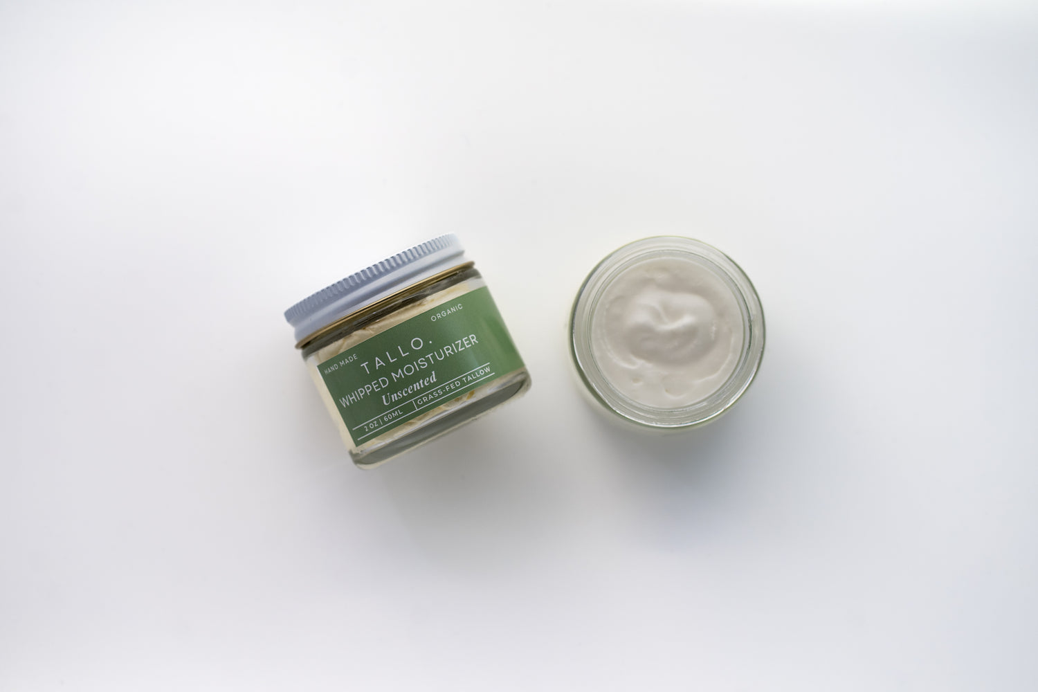 Two glass jars of Tallo Whipped Moisturizer with green labels and white lids, placed side by side. One jar is labeled 'Unscented,' while the other features a different variation. The labels indicate that the product is handmade, organic, and made from grass-fed tallow.