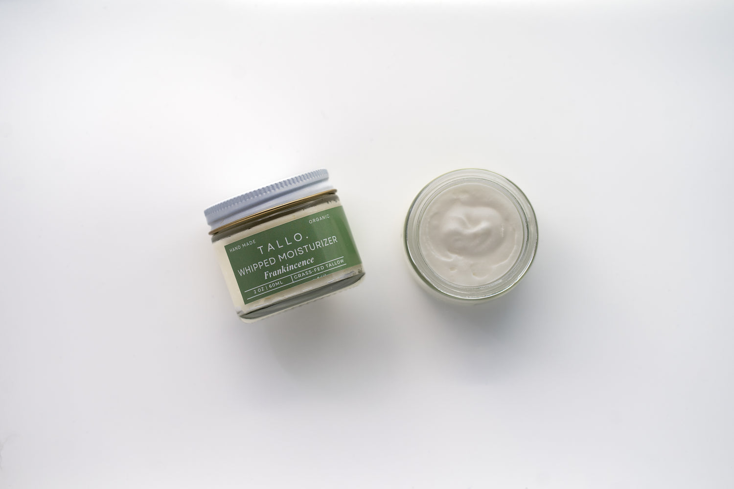 Two glass jars of Tallo Whipped Moisturizer with green labels and white lids, placed side by side. One jar is labeled 'Frankincense,' while the other features a different variation. The labels indicate that the product is handmade, organic, and made from grass-fed tallow.