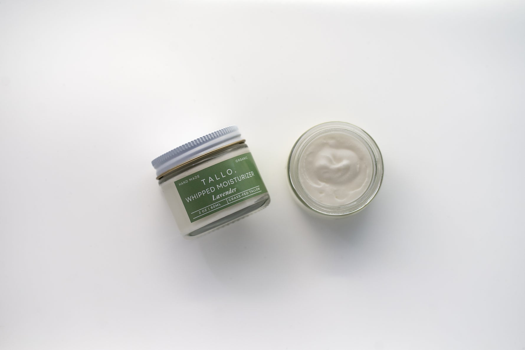 Two glass jars of Tallo Whipped Moisturizer with green labels and white lids, placed side by side. One jar is labeled 'Lavender,' while the other features a different variation. The labels indicate that the product is handmade, organic, and made from grass-fed tallow
