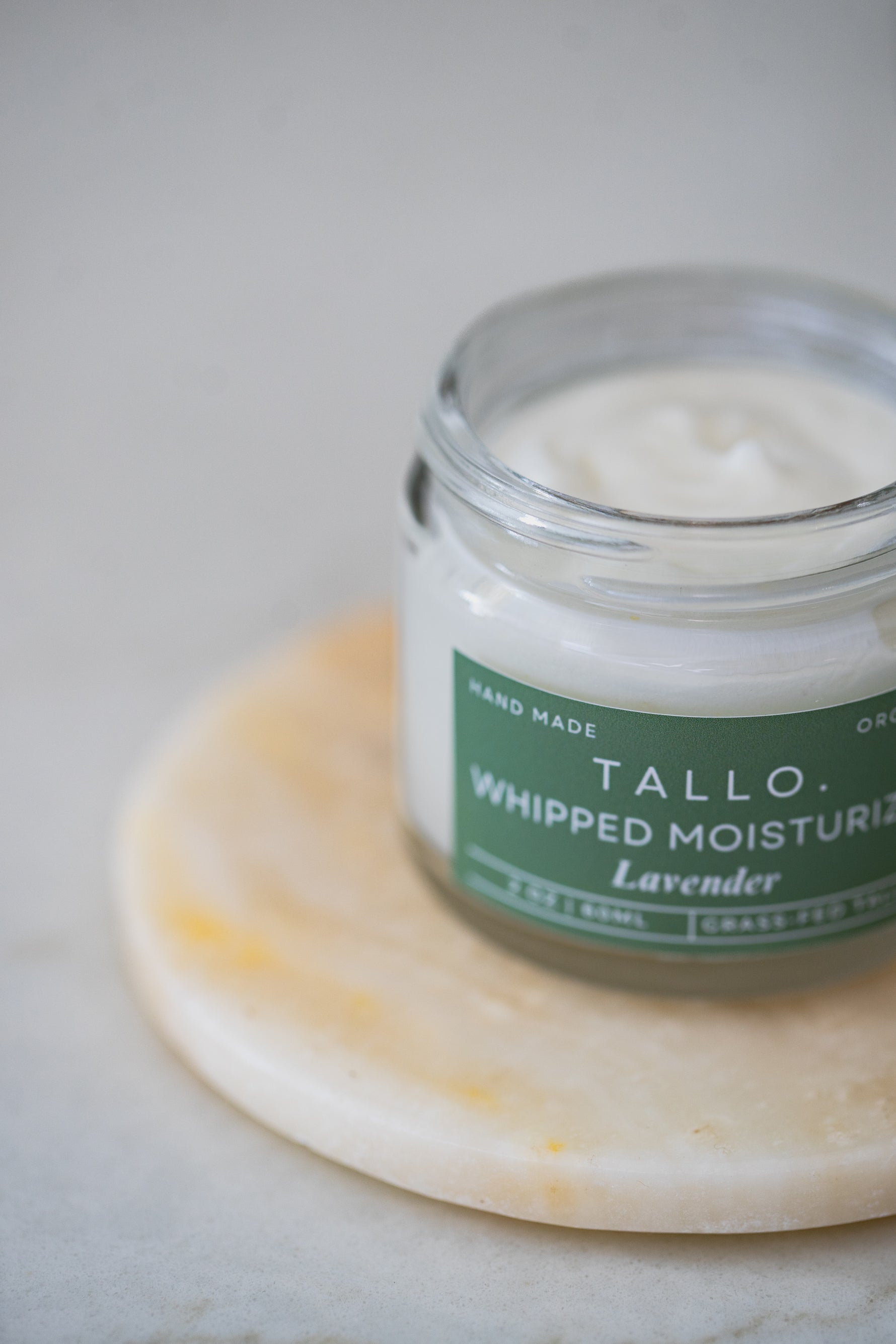 A 2 oz glass jars of Tallo Whipped Moisturizer with green labels and white lids, placed side by side. The jar is labeled 'Lavender,' sitting on a coaster. The labels indicate that the product is handmade, organic, and made from grass-fed tallow