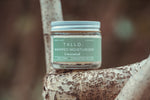 A glass jar of Tallo Whipped Moisturizer in the Unscented variation, placed on a textured tree branch. The jar has a green label with white text, indicating that the product is handmade, organic, and made from grass-fed tallow. The background is blurred with earthy tones.