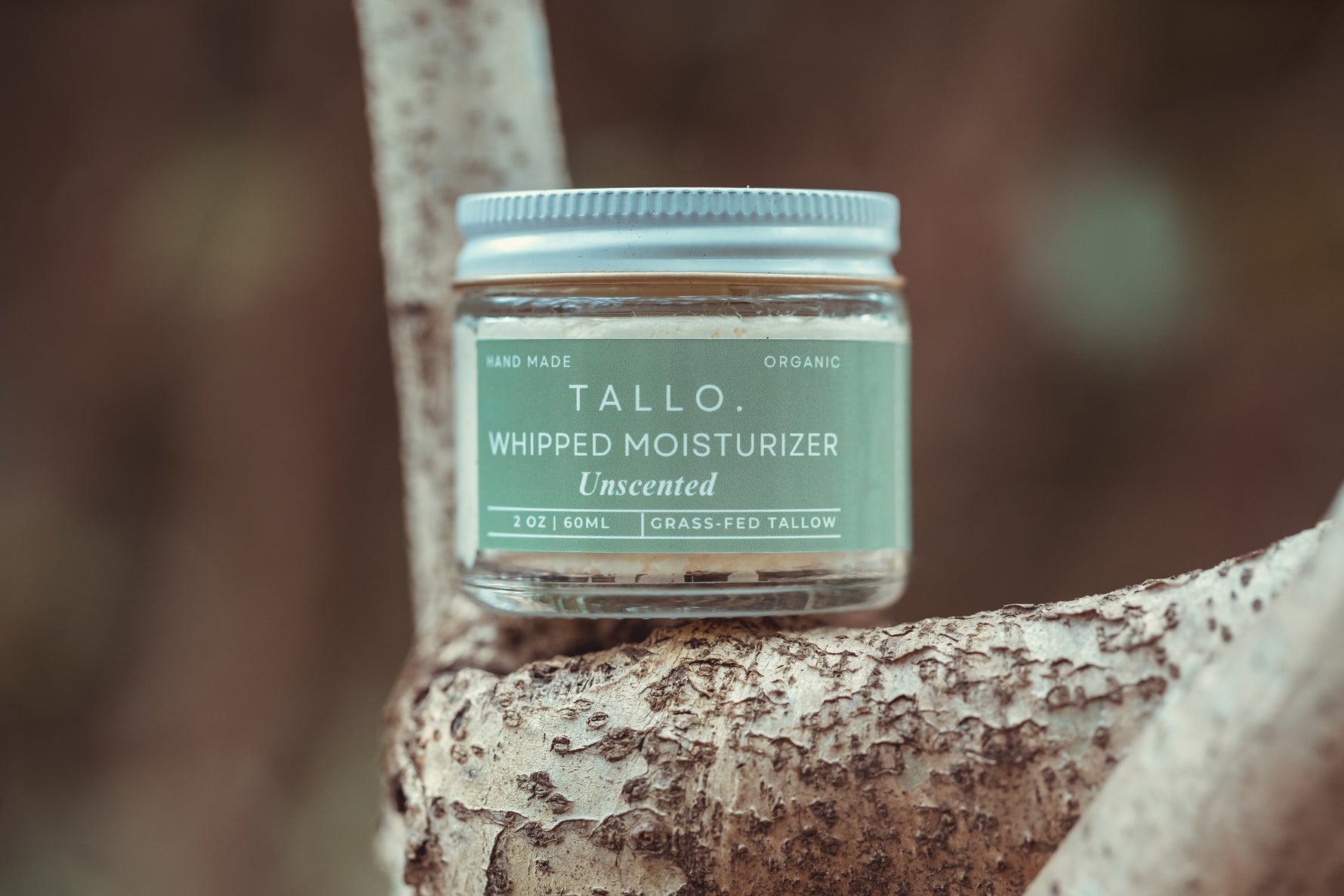 A glass jar of Tallo Whipped Moisturizer in the Unscented variation, placed on a textured tree branch. The jar has a green label with white text, indicating that the product is handmade, organic, and made from grass-fed tallow. The background is blurred with earthy tones.