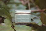 A glass jar of Tallo Whipped Moisturizer in the Unscented variation, placed among green leaves. The jar has a green label with white text, highlighting that the product is handmade, organic, and made from grass-fed tallow. The natural setting emphasizes an earthy, organic theme.