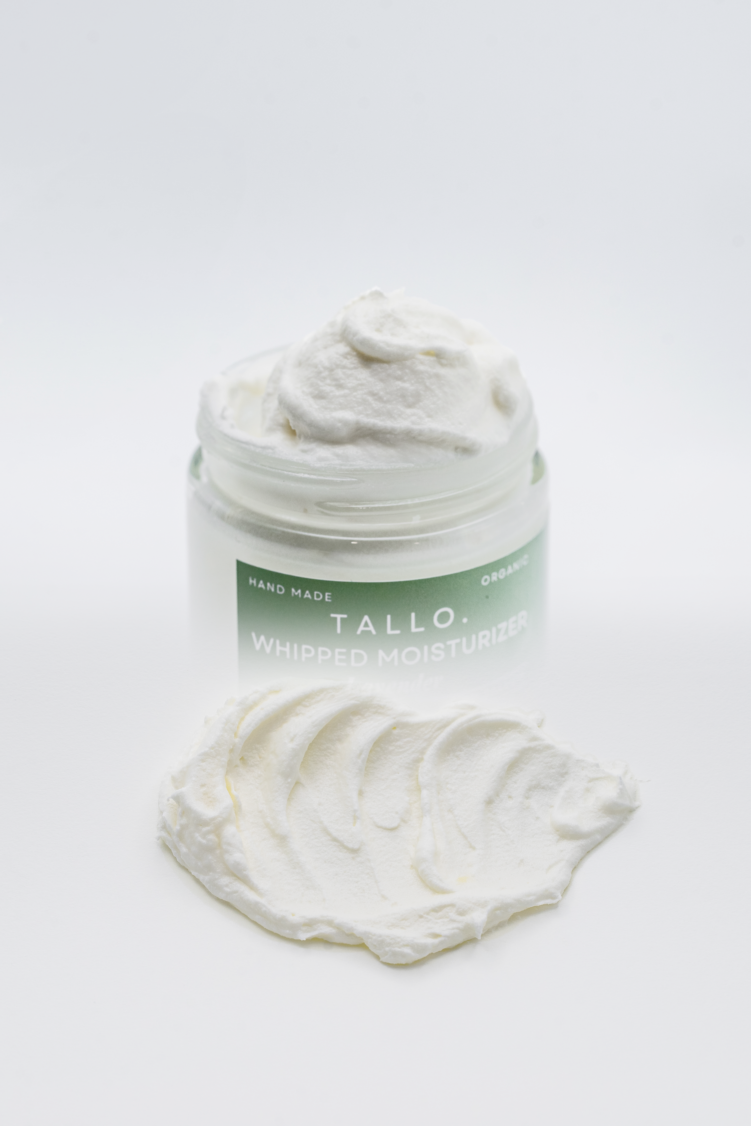 Gradient image of 2 oz Tallo Whipped Moisturizer with a green label. The 2nd gradient on the image shows the texture of the lotion being applied on a white background.