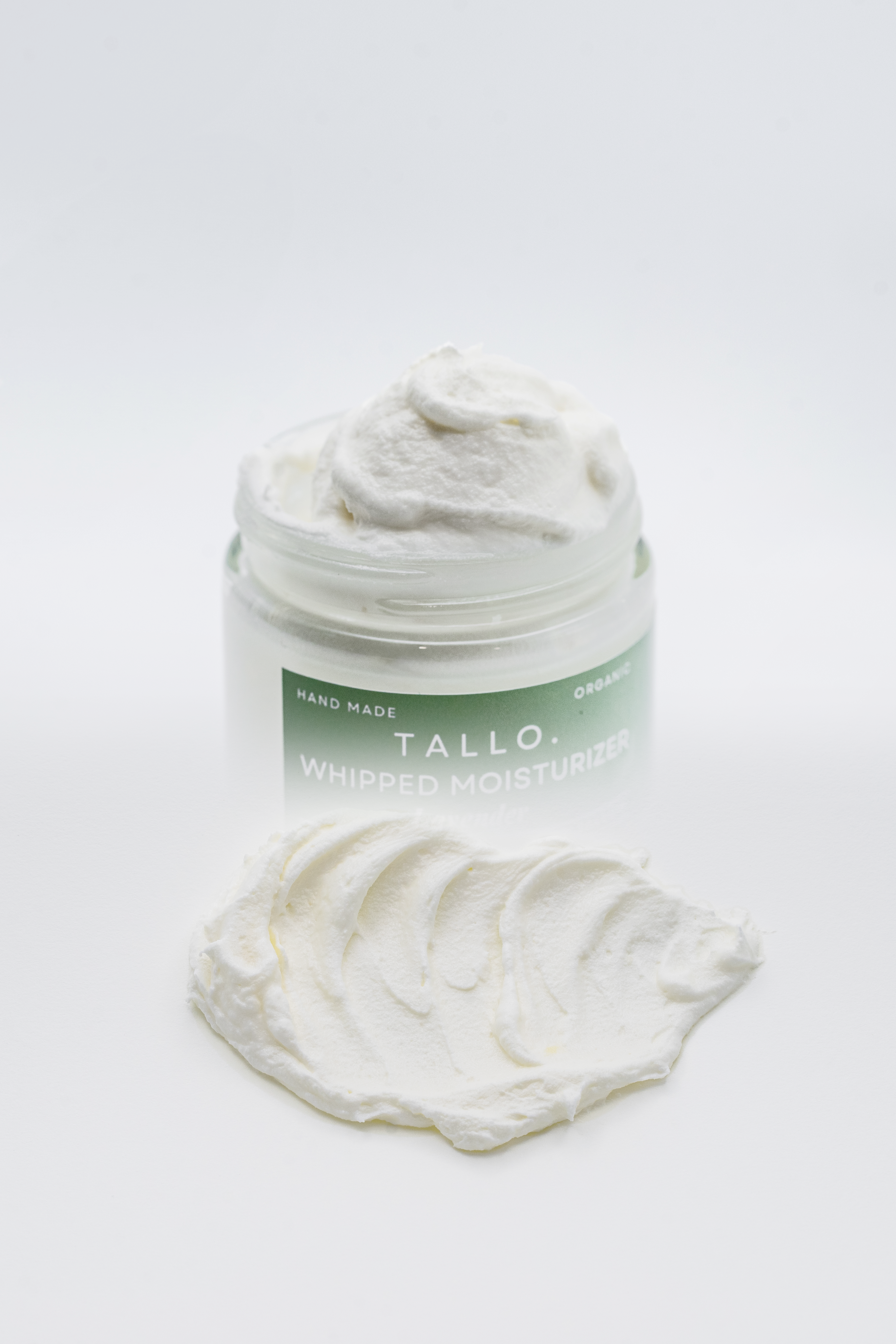 Gradient image of 2 oz Tallo Whipped Moisturizer with a green label. The 2nd gradient on the image shows the texture of the lotion being applied on a white background.